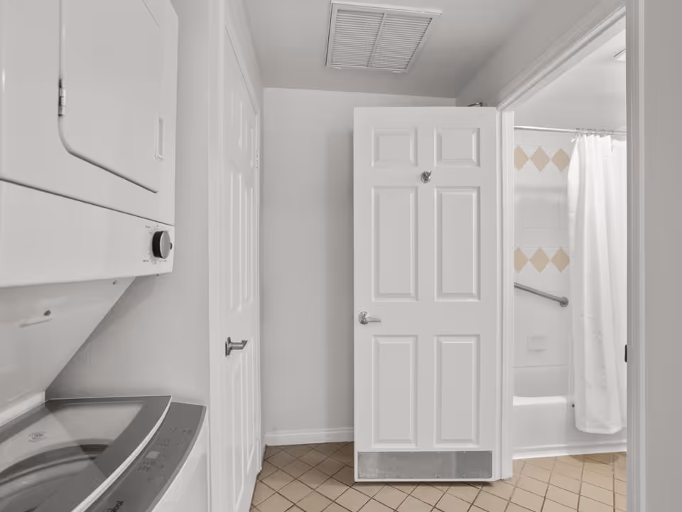 Laundry area with a stacked washer and dryer unit on the left side, adjacent to a white door that is partially open revealing a bathroom with a bathtub, shower curtain, and tiled walls with a diamond pattern.