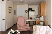 Cozy room with a pink wingback chair and floral sofa in front of a small kitchenette with a refrigerator and cabinets.