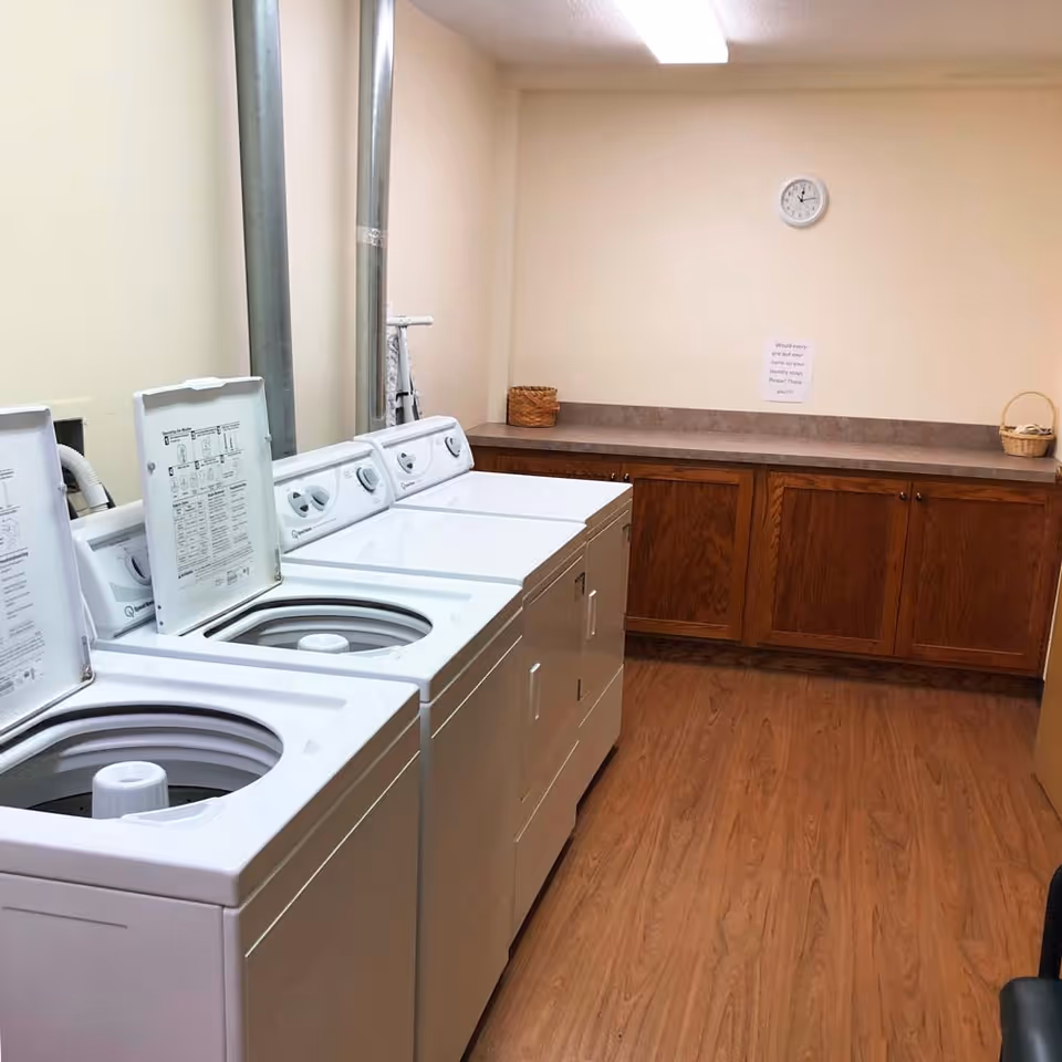 A clean communal laundry room with top-loading washing machines, a countertop with wooden cabinets, a wall clock, and small baskets.