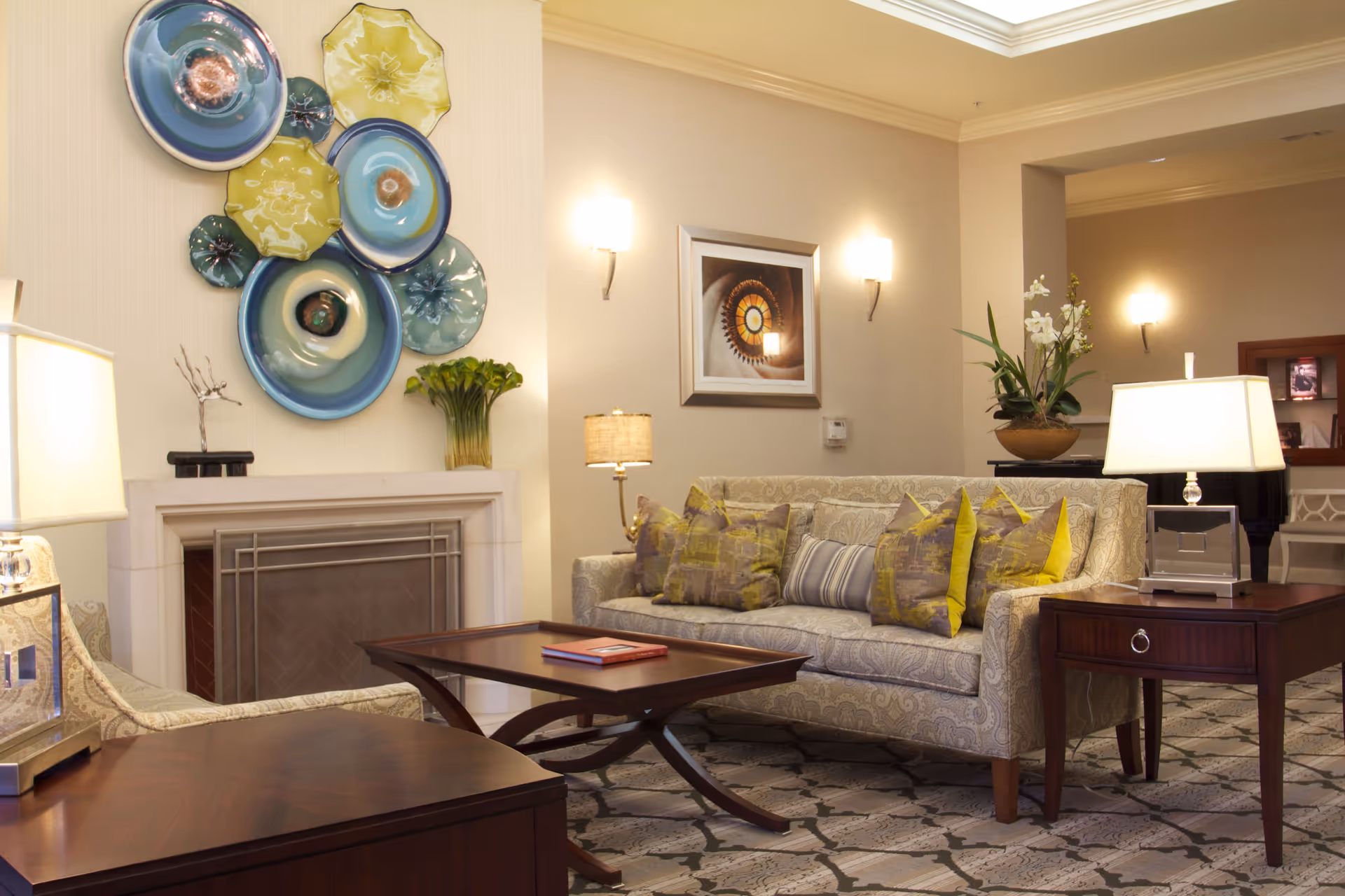 A cozy living room with a patterned beige sofa adorned with multiple yellow and gray pillows, a wooden coffee table with a book on it, and matching wooden side tables with lamps. The room features a fireplace with decorative ceramic plates mounted above it, a framed spiral staircase photo on the wall, and soft wall sconces providing warm lighting. A vase with flowers is placed on one of the side tables.