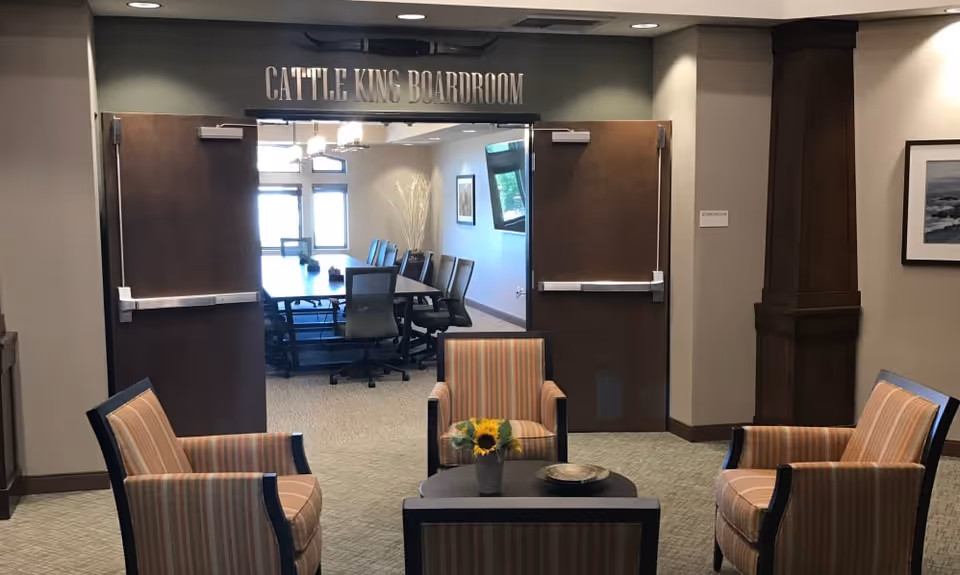 Interior view of a seating area with four striped armchairs arranged around a small round table with a sunflower centerpiece. In the background, double doors lead to a boardroom labeled 'Cattle King Boardroom' with a long table and office chairs inside.
