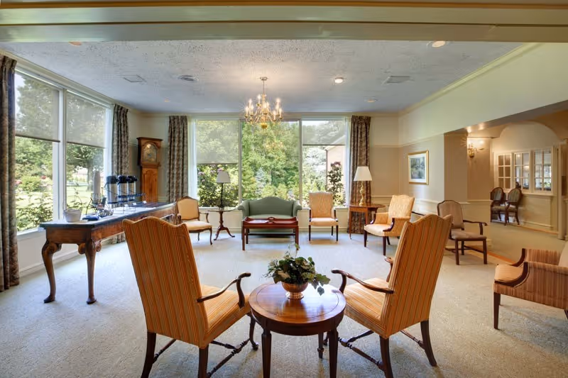 A bright and spacious living room area with large windows showing a green outdoor view. The room features multiple upholstered chairs arranged around small wooden tables, a grandfather clock, floor lamps, and a chandelier hanging from the ceiling. The decor is traditional with neutral tones and patterned curtains.