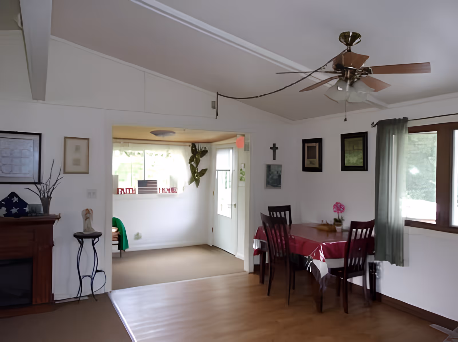 Interior view of a dining area with a wooden table covered by a red and white tablecloth, surrounded by four dark wooden chairs. The room has white walls with framed pictures and a cross hanging on one wall. A ceiling fan with lights is mounted on the ceiling. There is a window with green curtains letting in natural light. Adjacent to the dining area is another room with a window and a door, featuring a green jacket draped over a chair and a plant on the wall. A wooden mantel with decorative items is visible on the left side.