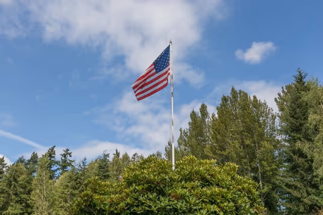 American flag flying on a flagpole above green bushes with tall trees and a partly cloudy blue sky in the background.