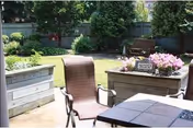 Outdoor patio area with a brown chair, raised garden beds with flowers, a wooden bench, and trees in the background under daylight.