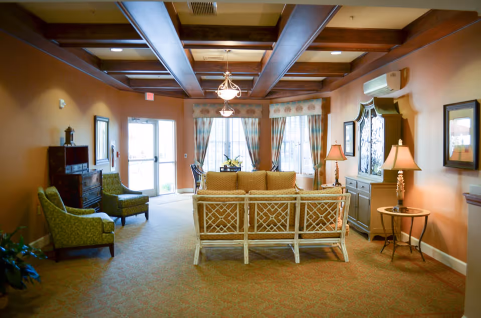 Warmly decorated lounge with sofas, armchairs, side tables and lamps beneath a beamed ceiling and windows near an entrance door.