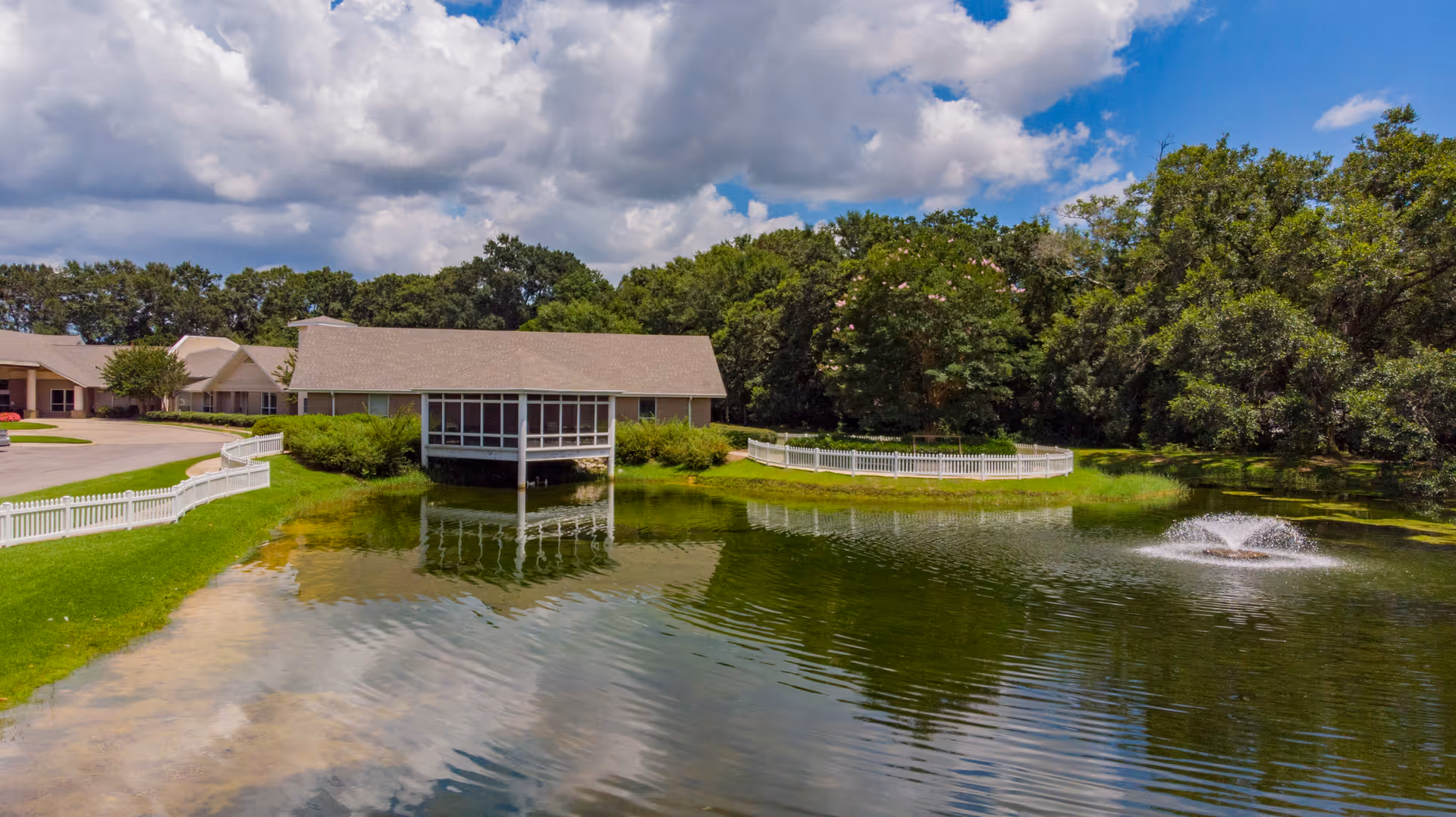 A serene outdoor scene at Live Oak Village featuring a pond with a small fountain, surrounded by green grass and trees. A building with a screened porch extends over the pond, and a white picket fence borders part of the grassy area near the water. The sky is partly cloudy with patches of blue.