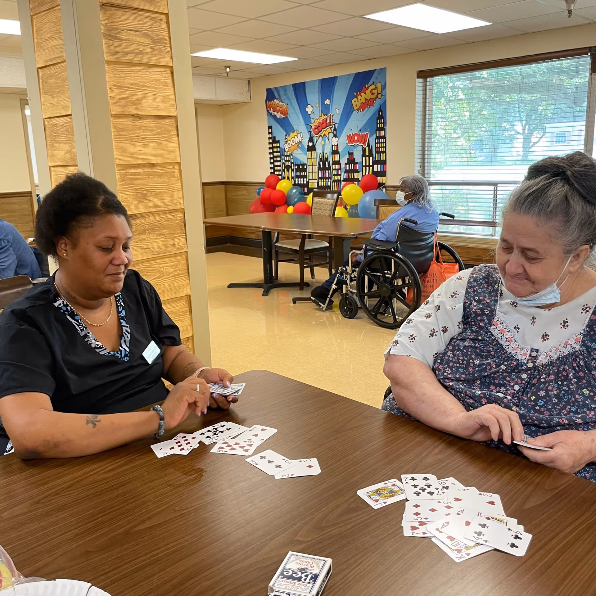 Two women play cards at a table in a communal activity/dining room while another resident in a wheelchair sits in the background.