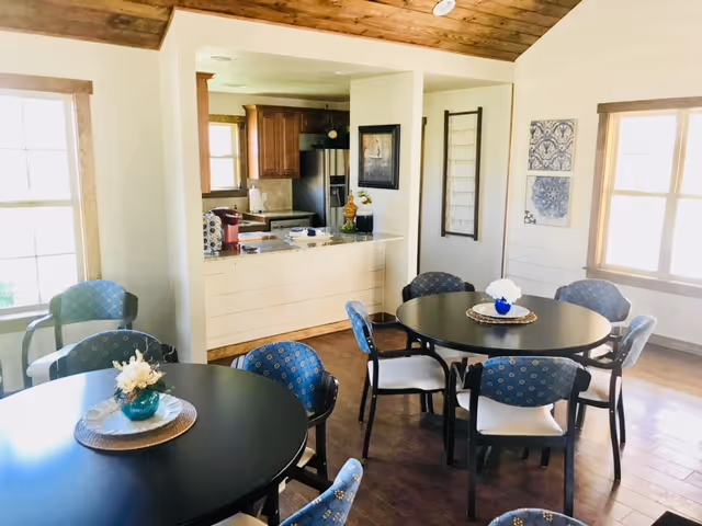 Bright communal dining area with round black tables and blue upholstered chairs beside an open kitchen and wood-paneled ceiling.