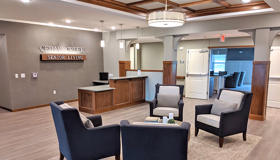 A modern senior living facility lobby with a reception desk, four cushioned armchairs arranged around a small table with books and decorative items, wood accents, and a sign on the wall that reads 'Cedar Creek Senior Living'.