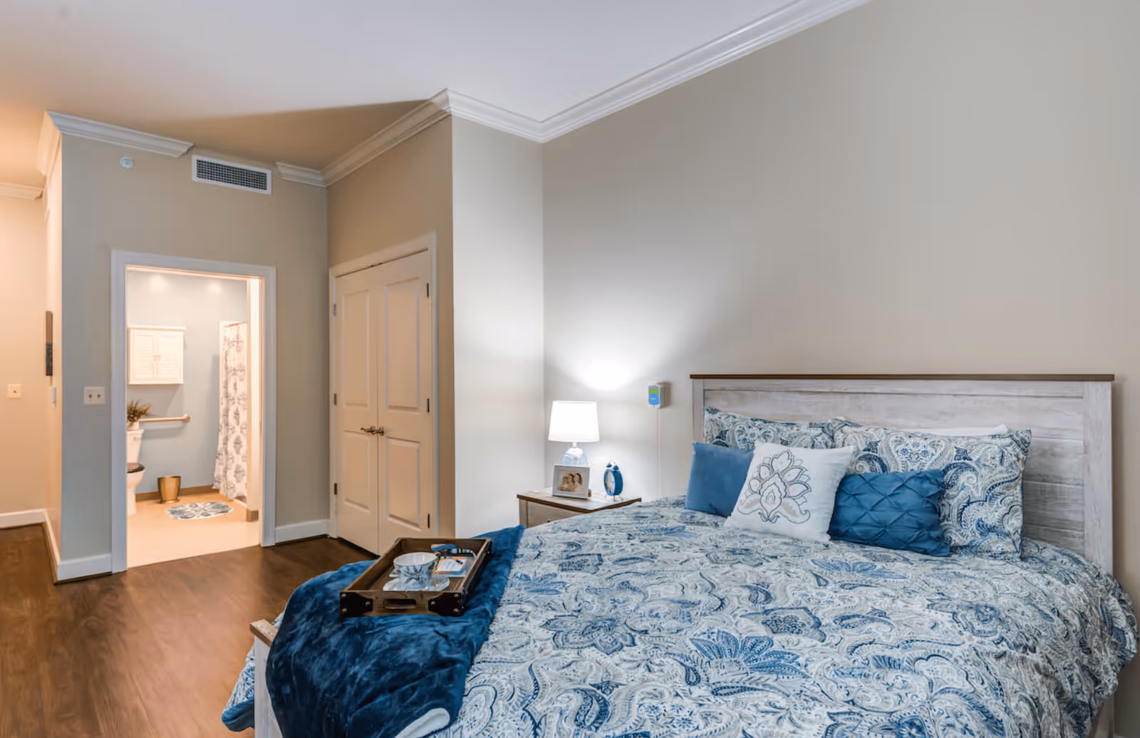 A cozy bedroom with a large bed featuring blue and white patterned bedding and multiple pillows. A wooden tray with a cup and saucer sits on a blue throw blanket at the foot of the bed. To the right of the bed is a nightstand with a lamp, a framed photo, and a small clock. The room has beige walls and wood flooring. An open doorway reveals a bathroom with a toilet, a shower curtain, and a small cabinet on the wall. There is also a closed double-door closet next to the bathroom entrance.