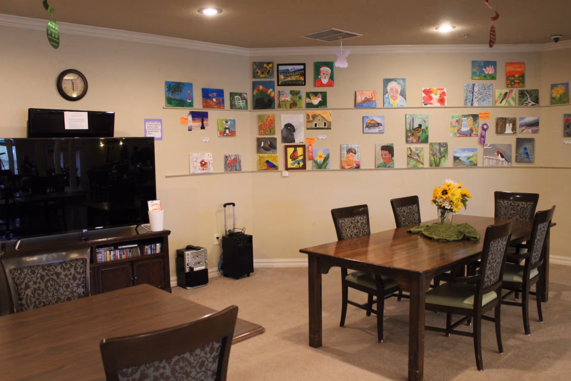 Community dining/activity room with a wooden table and chairs, a large TV, and colorful artwork displayed on the wall.