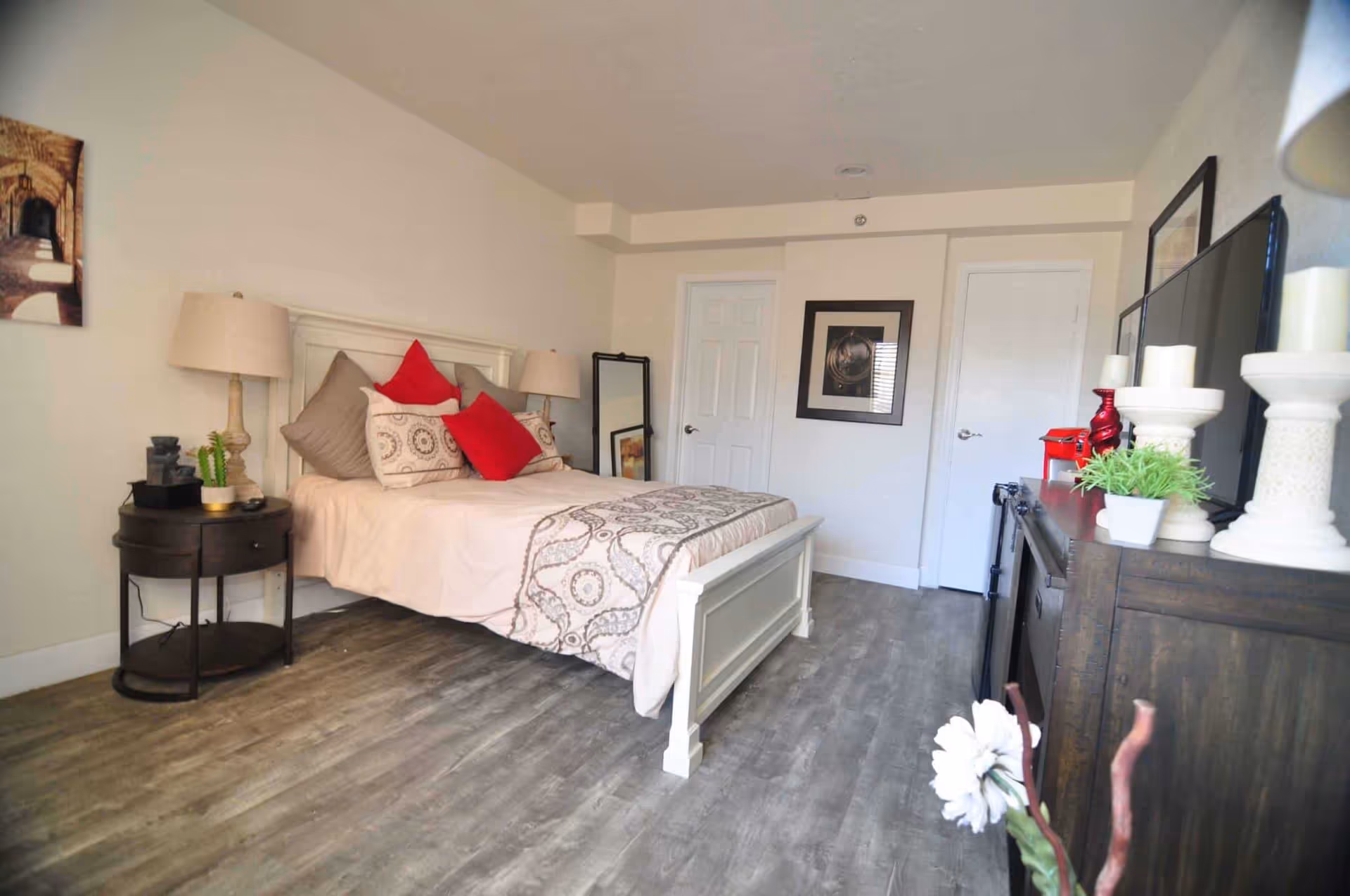 A bedroom with a white wooden bed frame, beige and patterned bedding, and red accent pillows. There are two bedside tables with lamps, a standing mirror, framed artwork on the walls, and a dark wooden dresser with decorative items including candles and a small plant. The floor is a light wood laminate.