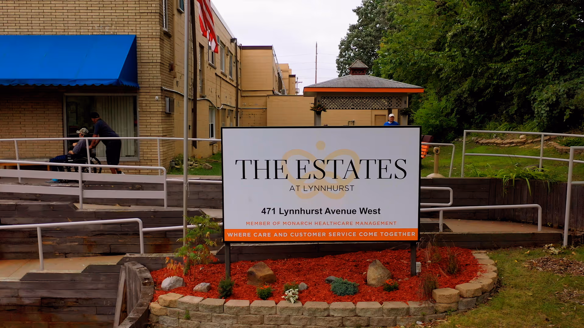 Outdoor view of The Estates at Lynnhurst senior living facility sign with landscaping around it, a building with a blue awning, an American flag, and two people near a wheelchair ramp in the background.