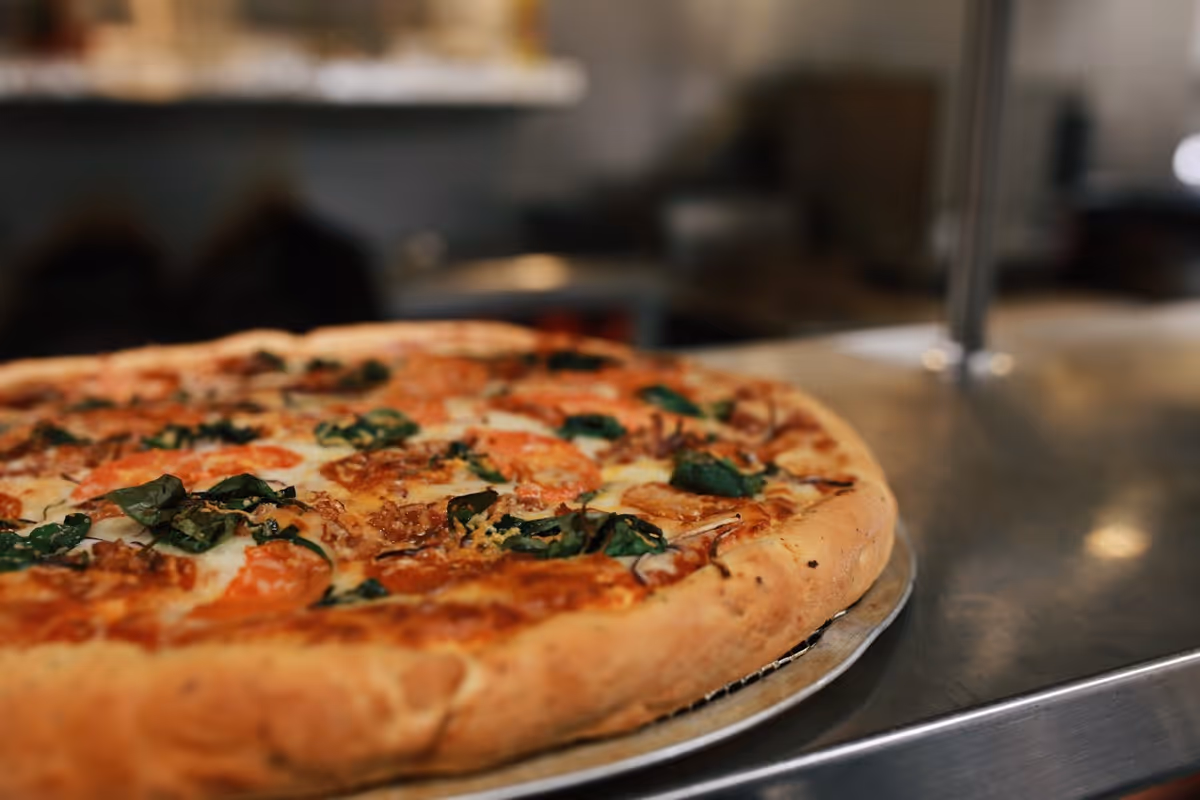 Large pizza topped with tomatoes, spinach, and melted cheese on a metal tray on a stainless-steel counter.