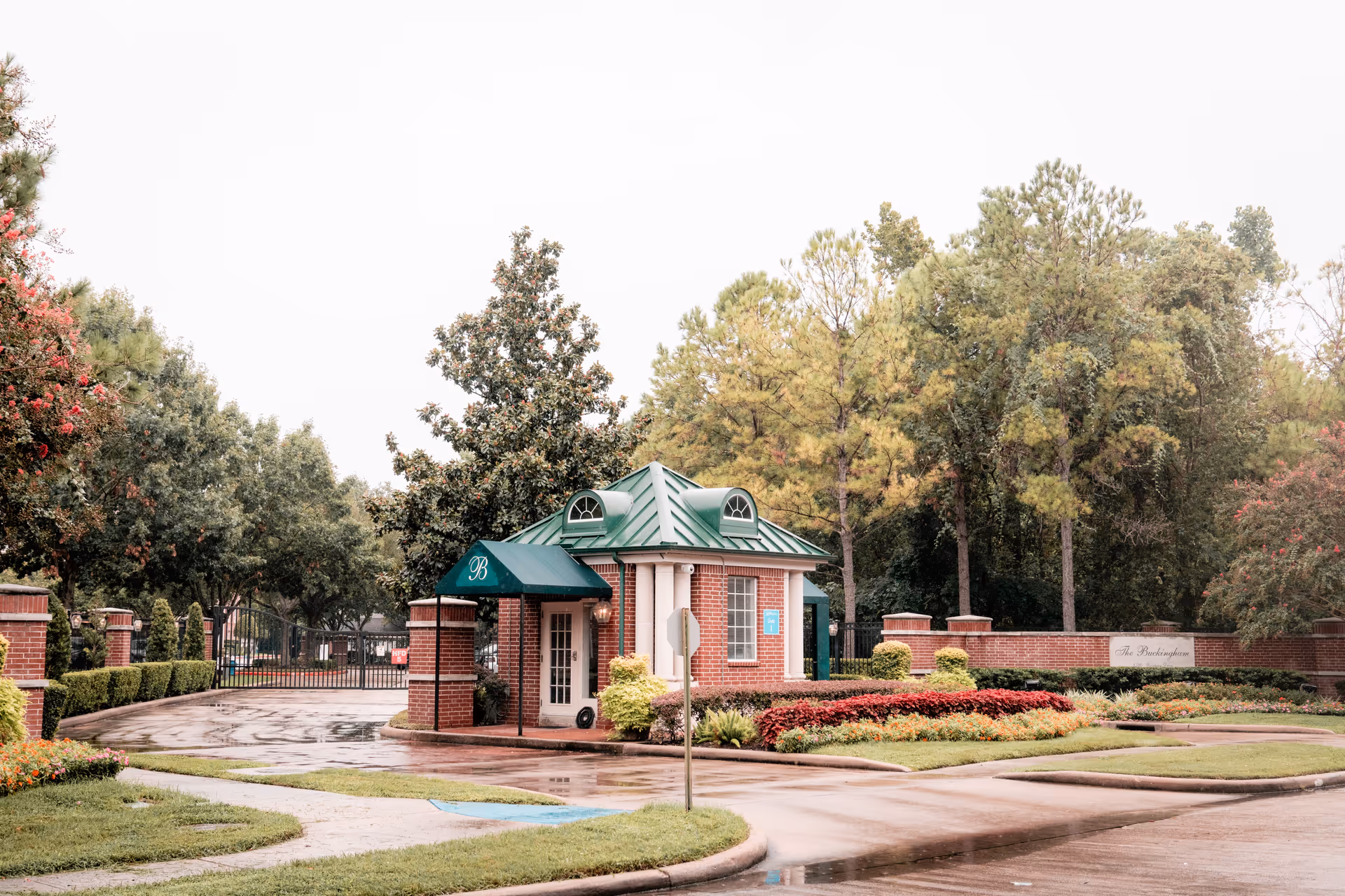 Entrance gatehouse of The Buckingham facility with a small brick building featuring a green metal roof and an awning with the letter B. The area is surrounded by trees, shrubs, and well-maintained flower beds. A gated entrance is visible behind the building.