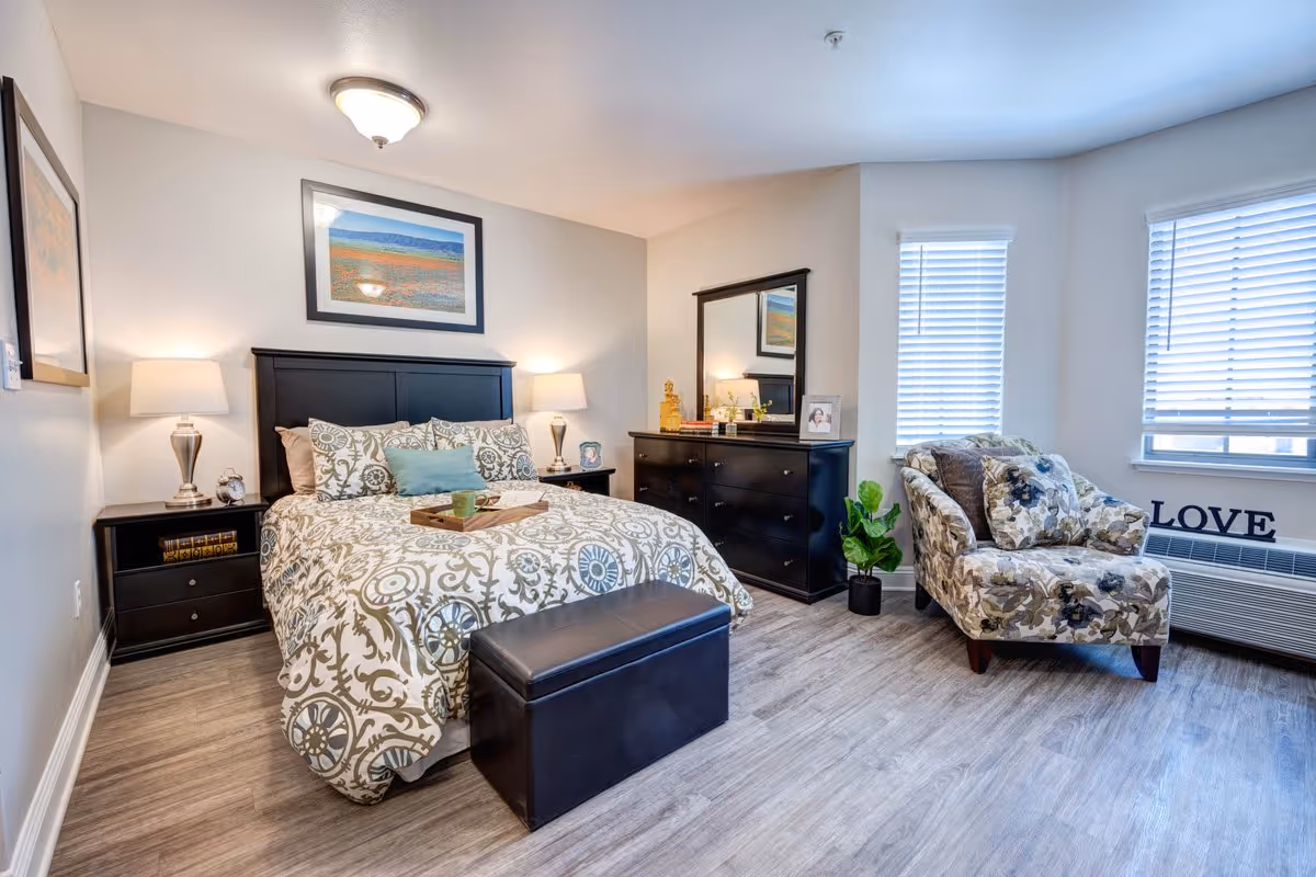A well-lit bedroom with a large bed featuring patterned bedding and multiple pillows. There are two bedside tables with lamps on either side of the bed. A black dresser with a mirror and decorative items is against the wall. A floral upholstered armchair is placed near two windows with blinds, and a small plant is next to the dresser. The word 'LOVE' is displayed on the windowsill above a heating/cooling unit. The floor is covered with light wood-style flooring.