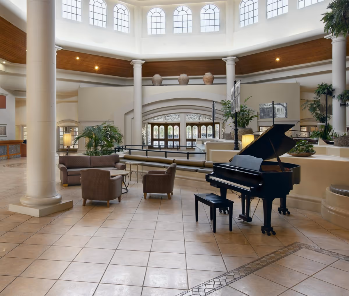 Spacious and elegant common area with a grand piano, seating arrangement including a sofa and two armchairs around a small table, large columns, potted plants, and tall arched windows letting in natural light.
