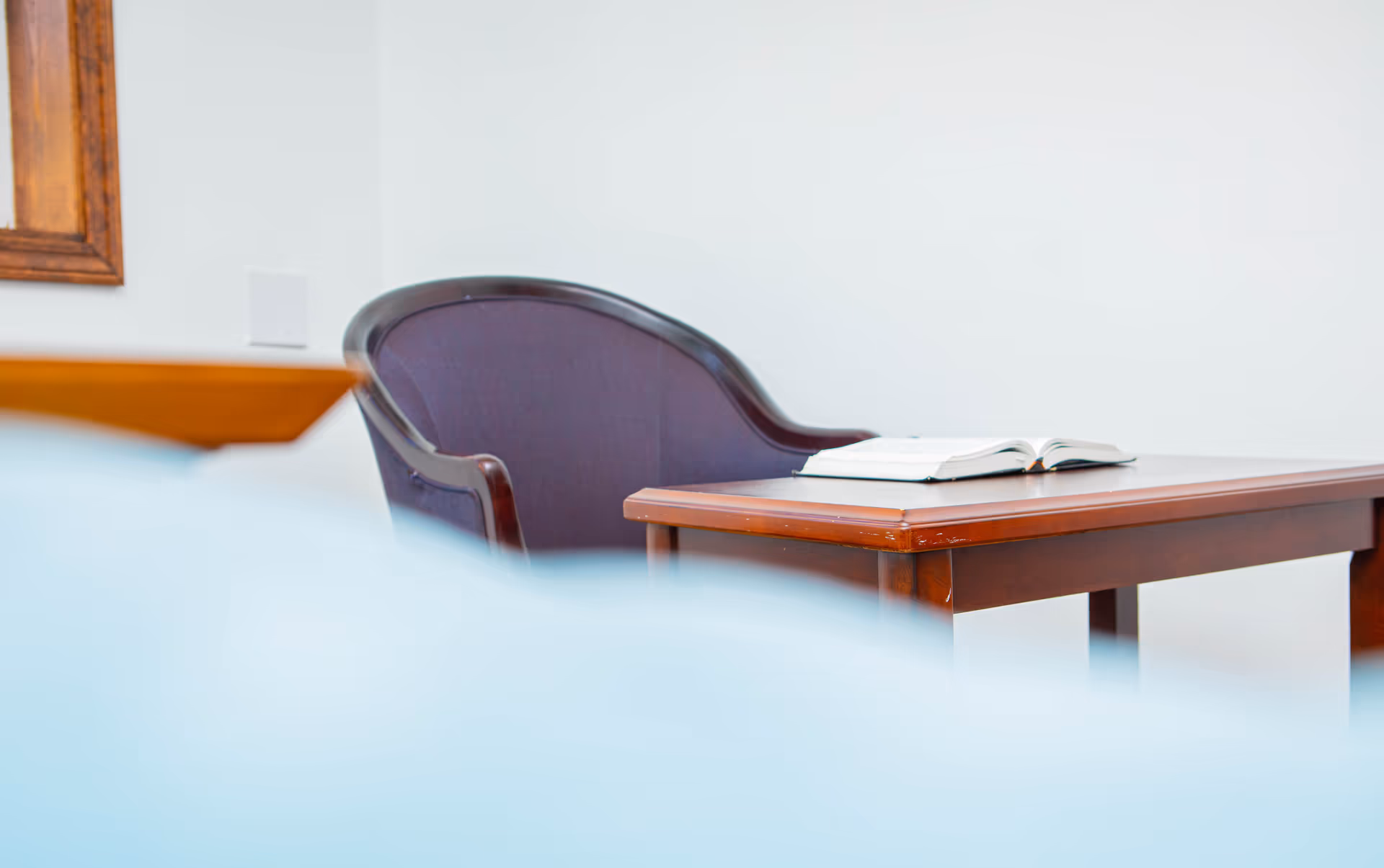 A wooden table with an open book on it, next to a dark upholstered chair, against a plain white wall with a wooden-framed mirror partially visible on the left.