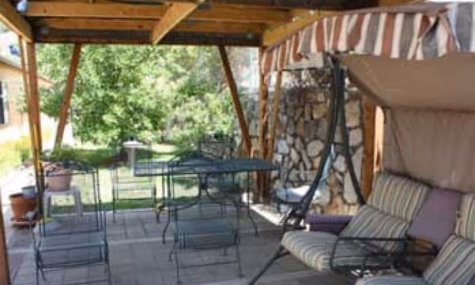 Covered outdoor patio area with metal table and chairs, a cushioned swing with striped canopy, potted plants, and a stone wall in the background with greenery beyond.