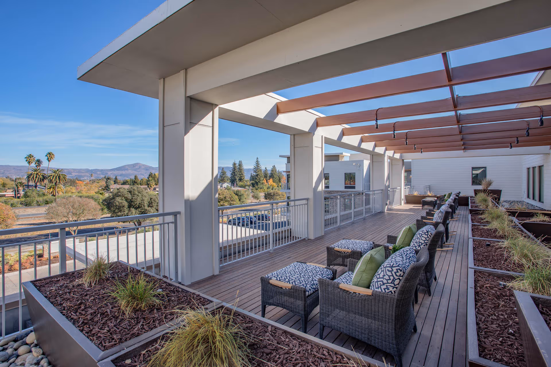 Outdoor terrace with wicker chairs and ottomans featuring patterned cushions and green pillows, surrounded by large planter boxes with plants. The terrace has a wooden deck floor and a pergola overhead with hanging lights. In the background, there is a scenic view of trees, palm trees, and distant mountains under a clear blue sky.