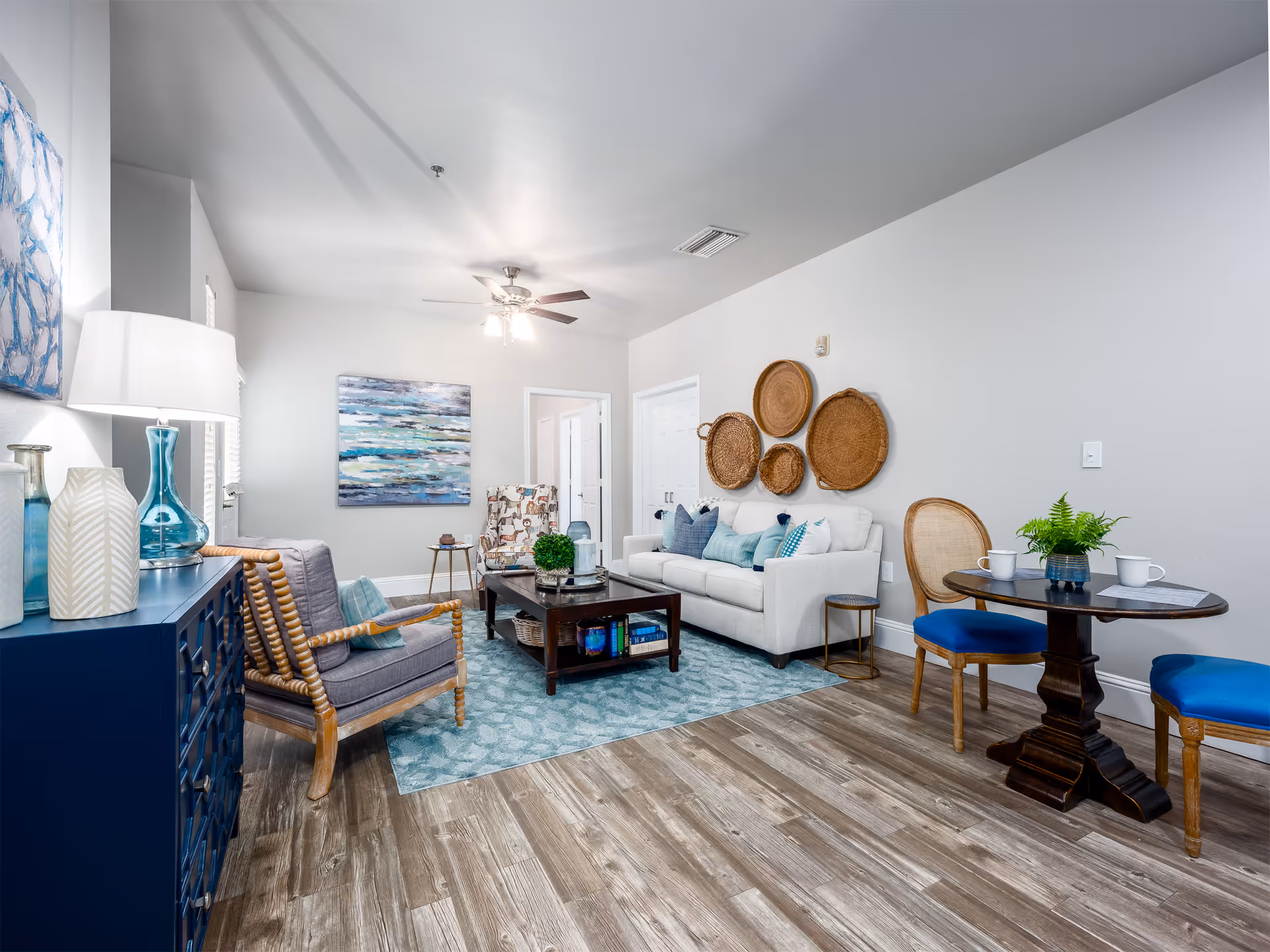 A bright and modern living room with light gray walls and wood flooring. The room features a white sofa adorned with blue and teal pillows, a wooden coffee table with decorative items, and two armchairs with wooden frames and gray cushions. On the wall above the sofa, there are four woven basket-style wall decorations. A ceiling fan with lights is mounted on the ceiling. To the right, there is a small round wooden table with two blue cushioned chairs and a small plant on the table. On the left side, a blue cabinet holds decorative vases and a lamp. A large abstract painting hangs on the far wall.