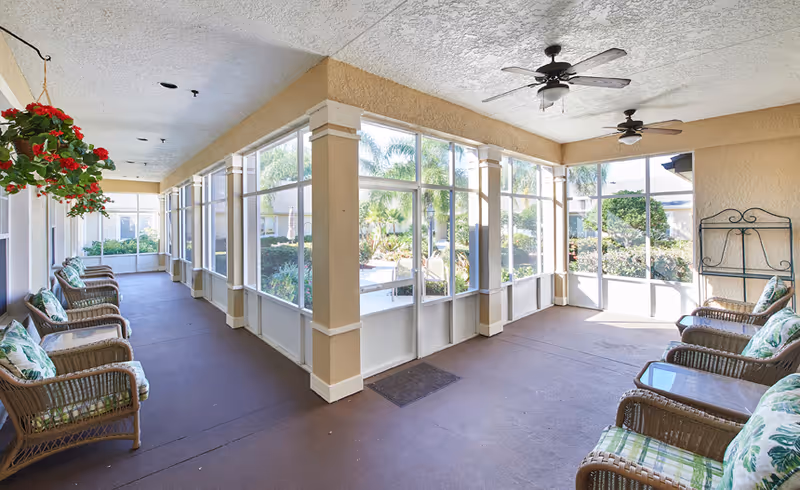 A spacious screened-in porch area with wicker chairs arranged along the walls, each with green and white patterned cushions. The porch has ceiling fans and large windows providing a view of a garden with palm trees and other greenery outside.
