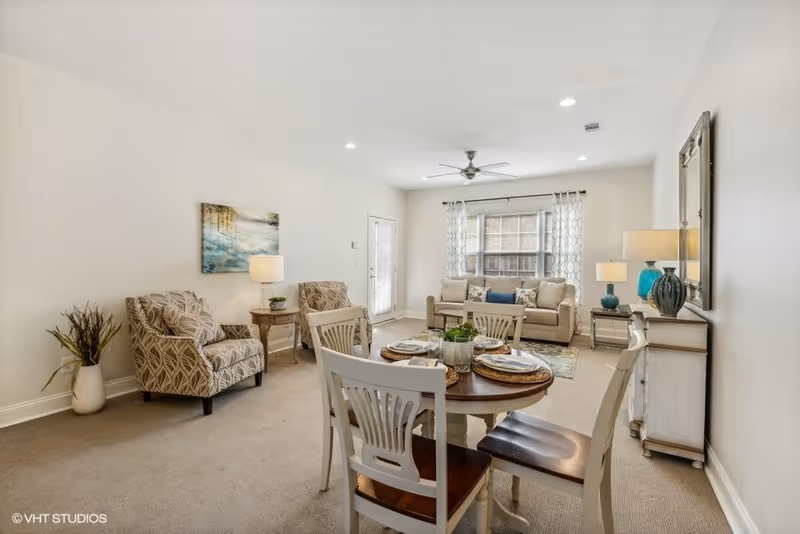 A bright and cozy living room and dining area with a round wooden dining table set for four, two patterned armchairs, a beige sofa with cushions, a ceiling fan, and decorative lamps and artwork on the walls.