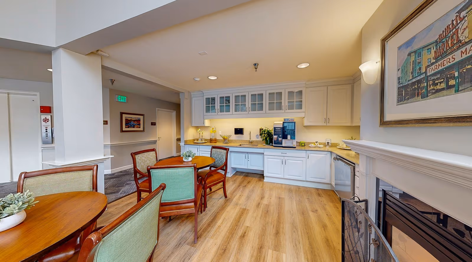 Bright communal dining and kitchenette area with round wooden tables and upholstered chairs, white cabinets, and a fireplace.