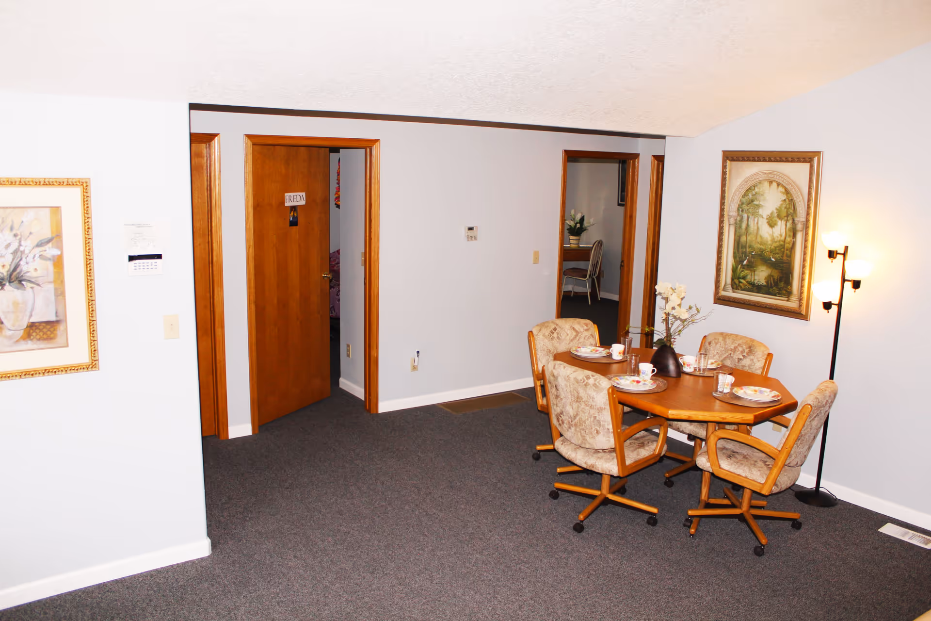 Small interior dining area with a round wooden table set for four surrounded by upholstered rolling chairs, framed artwork, a floor lamp, and open doorways.