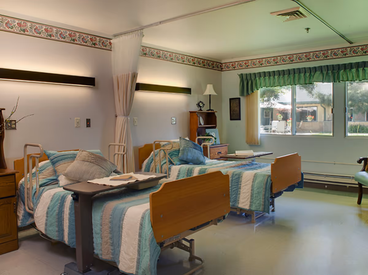 A room with two hospital-style beds, each made with blue and white striped bedding and pillows. There is a curtain between the beds for privacy, a wooden nightstand with a lamp and decorative items, and a large window with green curtains letting in natural light. The room has a clean and calm atmosphere.