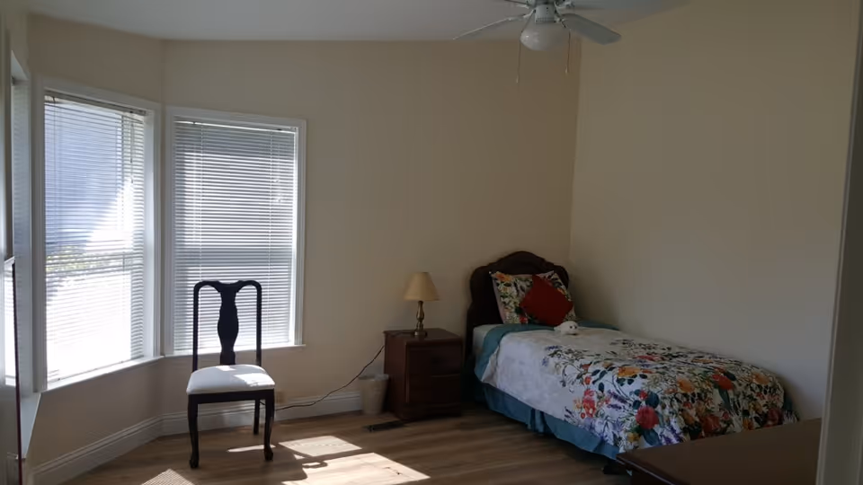 A simple bedroom with a single bed covered in floral bedding, a wooden nightstand with a lamp, a wooden chair near a bay window with blinds, and a ceiling fan above. The room has light-colored walls and wooden flooring.
