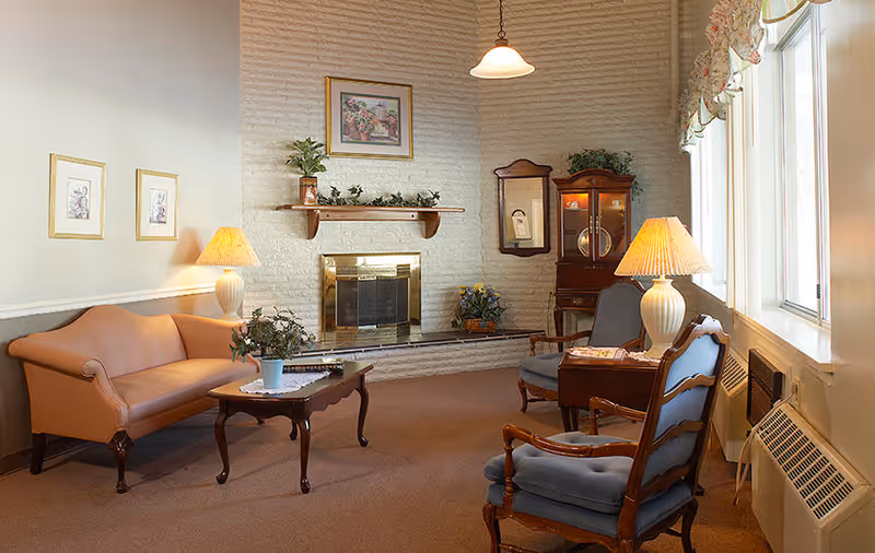 A cozy living room with a beige couch, two blue cushioned wooden armchairs, a wooden coffee table with a potted plant, two table lamps with beige shades, a white brick fireplace with a gold trim, a wooden cabinet with glass doors, a wall mirror, and framed artwork on the walls. The room has large windows with floral valances letting in natural light.