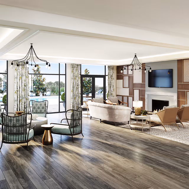 A spacious and bright living room area with large windows and glass doors leading to an outdoor pool area. The room features modern furniture including cushioned chairs, a sofa, a coffee table, and a fireplace with a mounted flat-screen TV above it. The floor is wooden, and the room is decorated with patterned curtains and contemporary light fixtures.