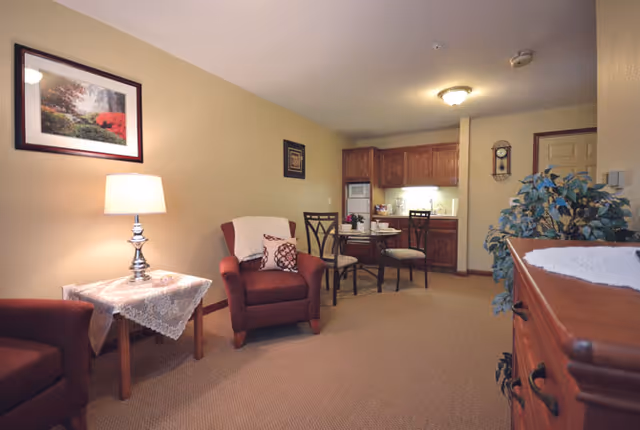 A cozy senior living room area with two red armchairs, a small wooden side table with a lace tablecloth and a lamp, a framed picture on the wall, a small dining table with two chairs, and a kitchenette with wooden cabinets in the background. There is also a wooden dresser with a plant on top and a wall clock near the door.