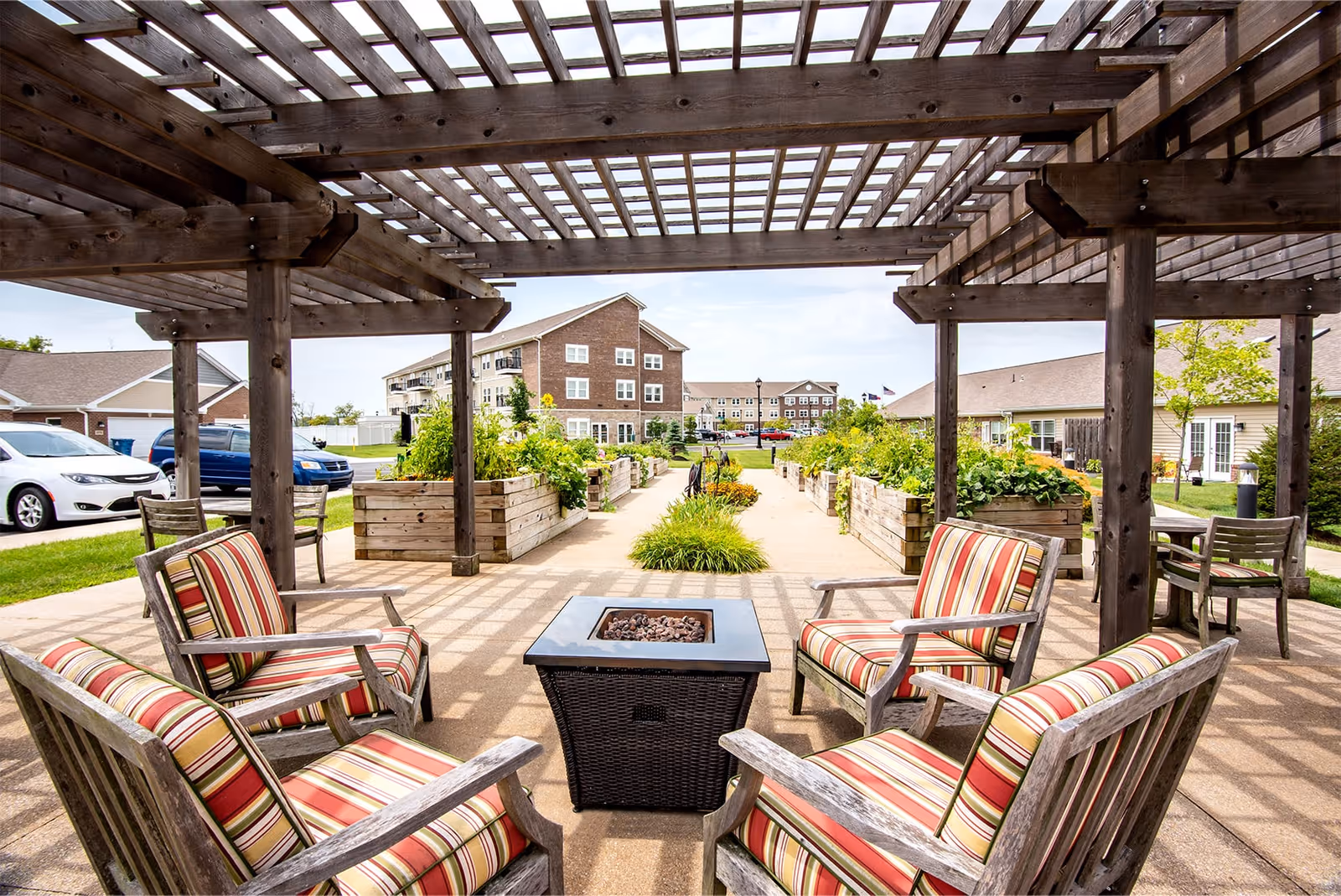 Outdoor communal patio with striped cushioned chairs arranged around a fire pit under a wooden pergola, with raised garden beds and buildings in the background.
