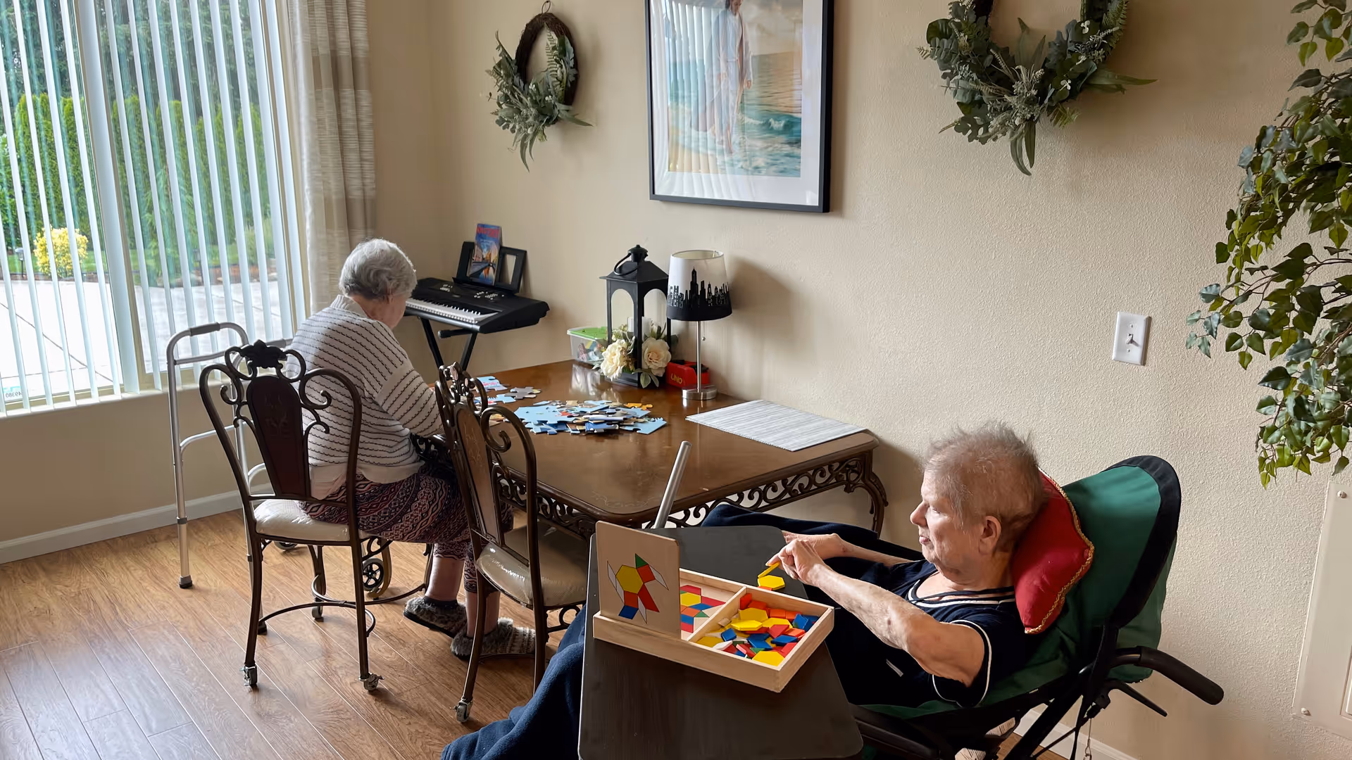 Two elderly residents seated at a table near a large window working on puzzles and shape-sorting activities in a cozy common room.