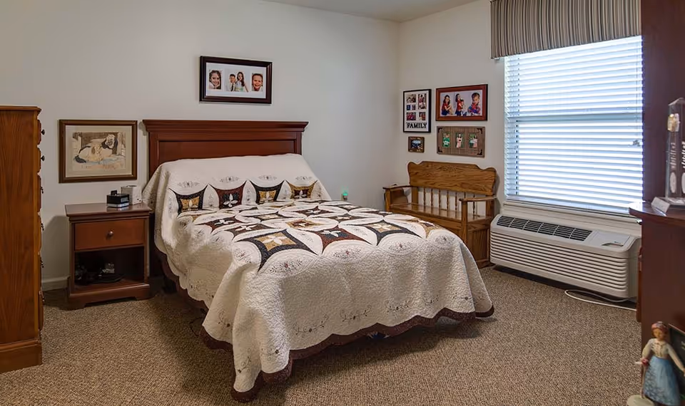 A cozy bedroom with a wooden bed frame and a quilted bedspread featuring a geometric pattern. There is a wooden nightstand with a drawer and an open shelf next to the bed. The walls are decorated with framed family photos and artwork. A wooden bench is placed against the wall near a window with closed blinds. The room has carpeted flooring and an air conditioning unit below the window.