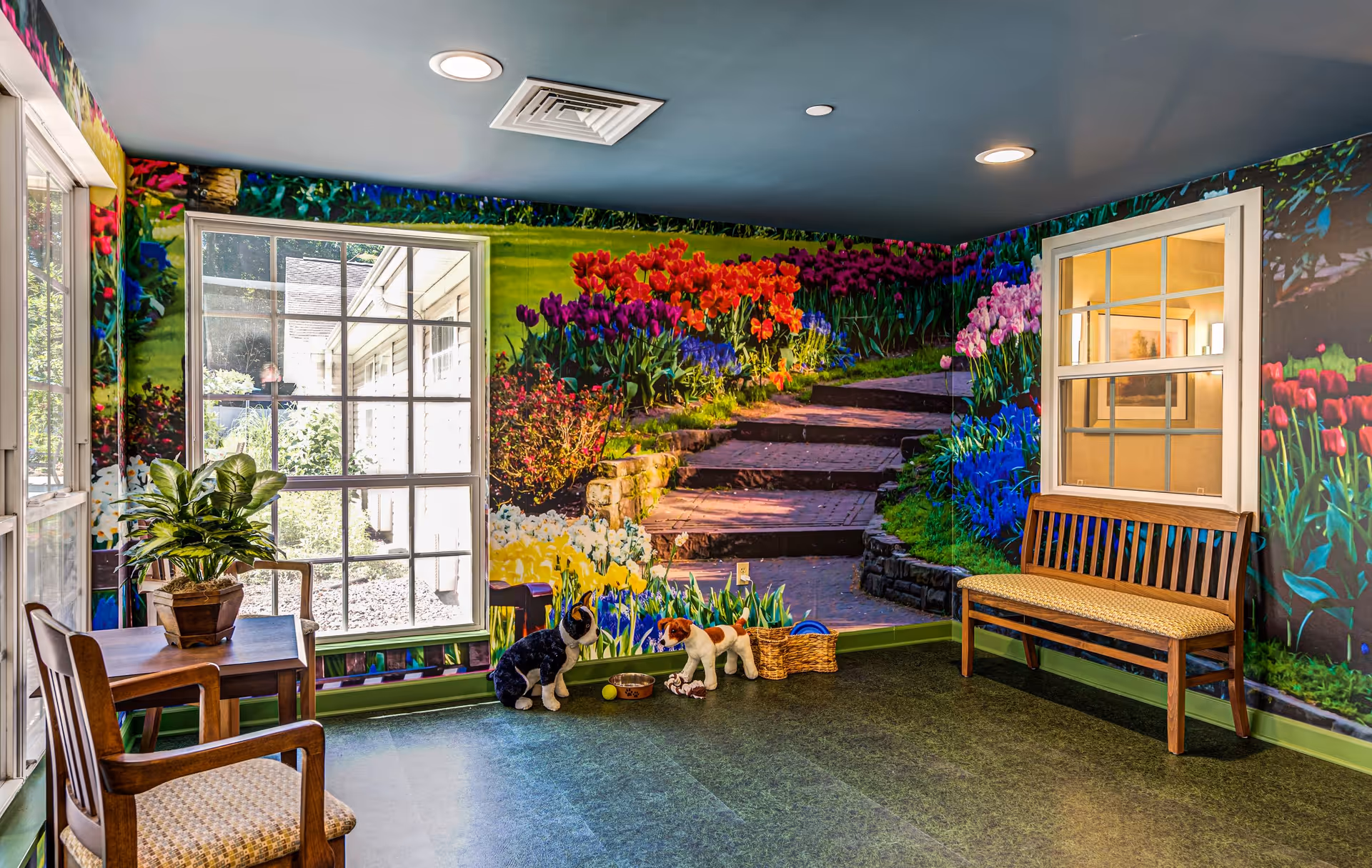 A bright and colorful interior room with a large mural of a garden pathway surrounded by vibrant flowers covering the walls. The room has a window looking outside, a wooden bench with a cushioned seat, two wooden chairs, a small table with a potted plant, and two dog statues with a basket and toys on the floor.