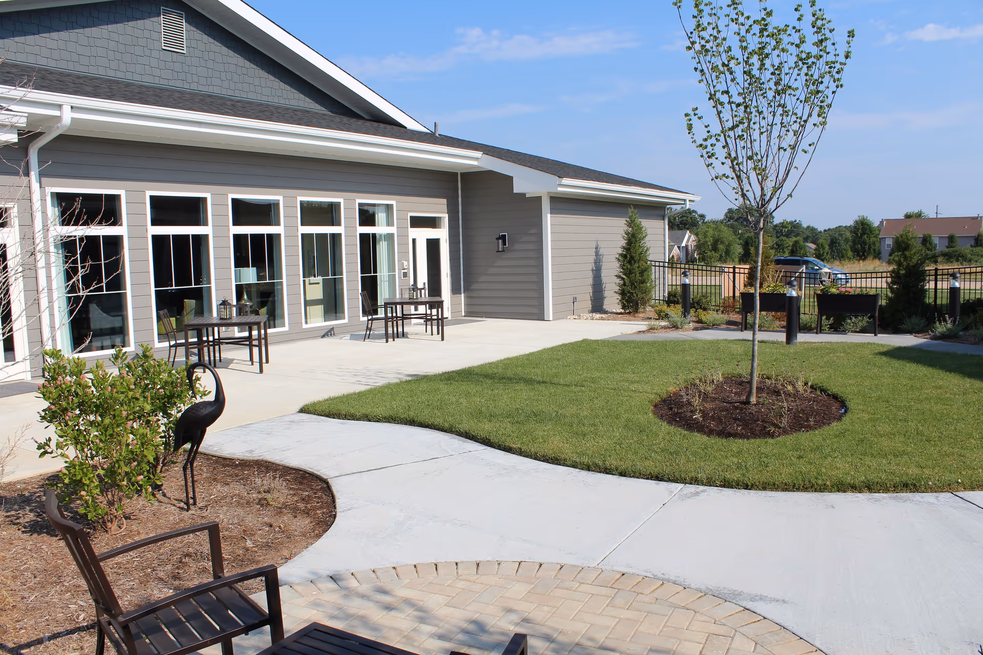 Outdoor patio area at Cottages of Lake St Louis featuring a concrete walkway, green lawn with a young tree, several small tables with chairs, and a decorative metal bird sculpture near some bushes. The building exterior is gray with large windows and a door, under a clear blue sky.