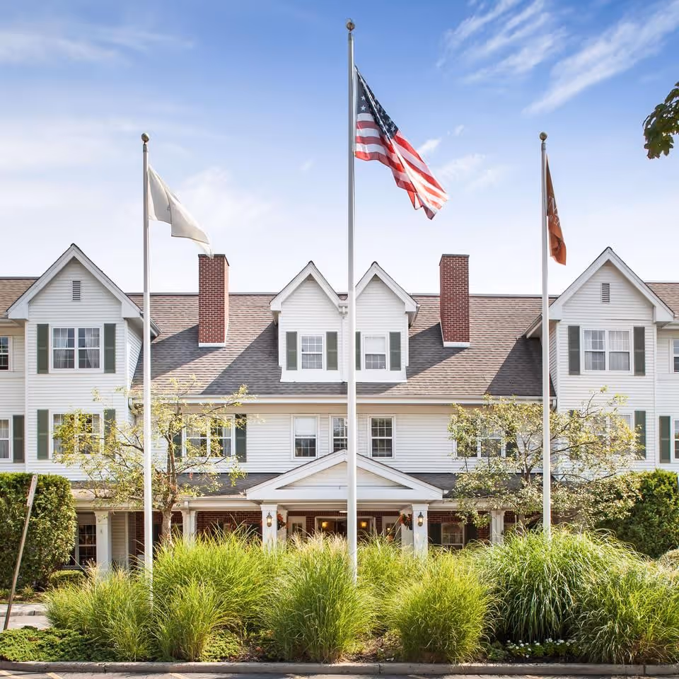 Exterior view of Brighton Gardens of Mountainside with flagpoles, shrubs, and a white facade.