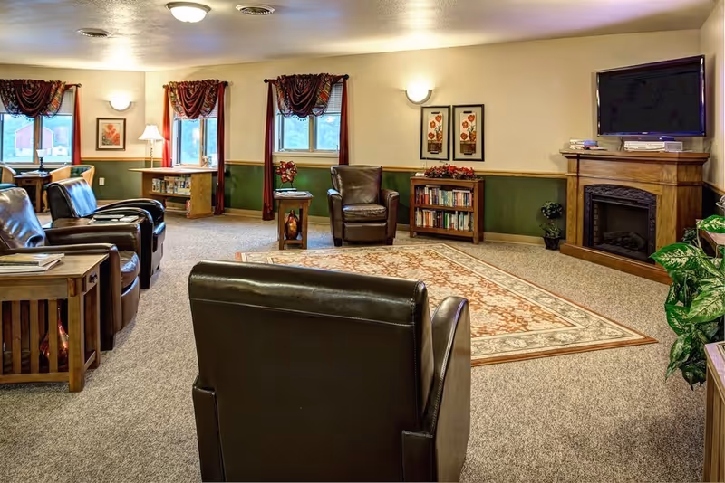 A cozy living room with several dark leather armchairs arranged around a patterned area rug. The room features a wooden fireplace with a flat-screen TV mounted above it, a small bookshelf with books and decorative items, and windows with red curtains letting in natural light. The walls are painted beige with a green wainscoting, and there are framed floral artworks on the walls.
