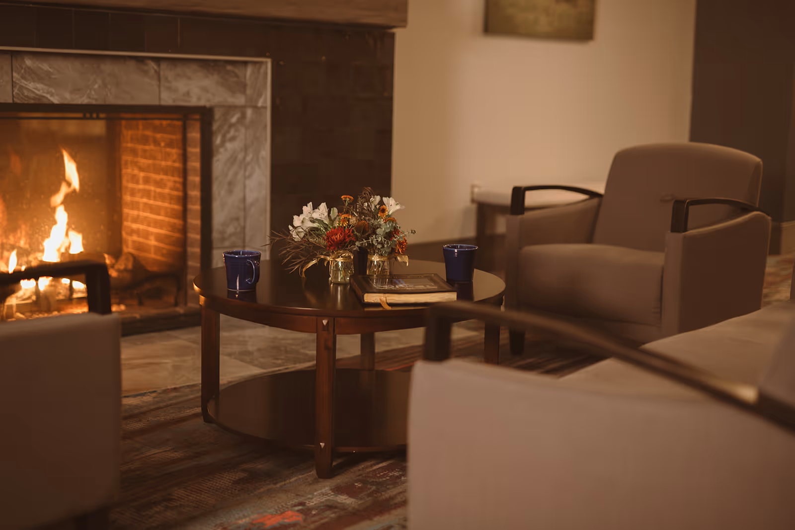 Cozy living room area with a lit fireplace, a round wooden coffee table with a floral arrangement, two blue mugs, and a book. Surrounding the table are comfortable beige armchairs with wooden armrests.