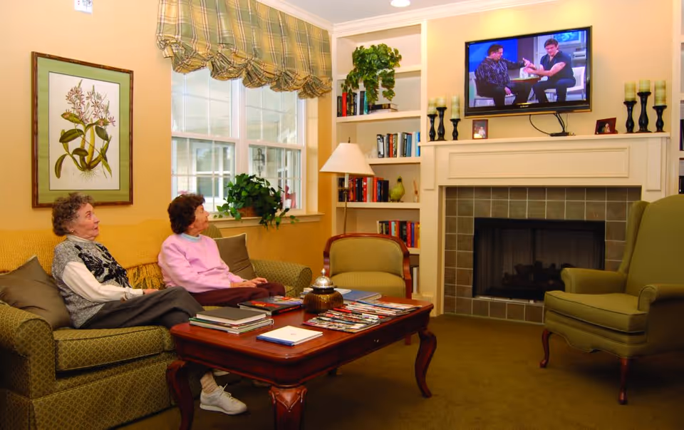 Two elderly women sitting on a green patterned couch in a cozy living room with a wooden coffee table in front of them. The room features a fireplace with a mounted TV above it, built-in bookshelves filled with books and decorative items, a green armchair, and a window with plaid valance curtains letting in natural light.