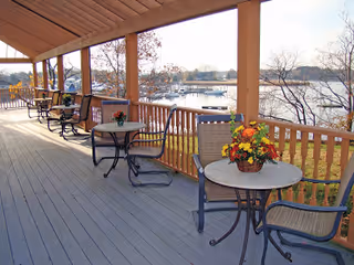 Covered outdoor patio with several round tables and chairs arranged along a wooden railing overlooking a body of water with boats and trees in the background.