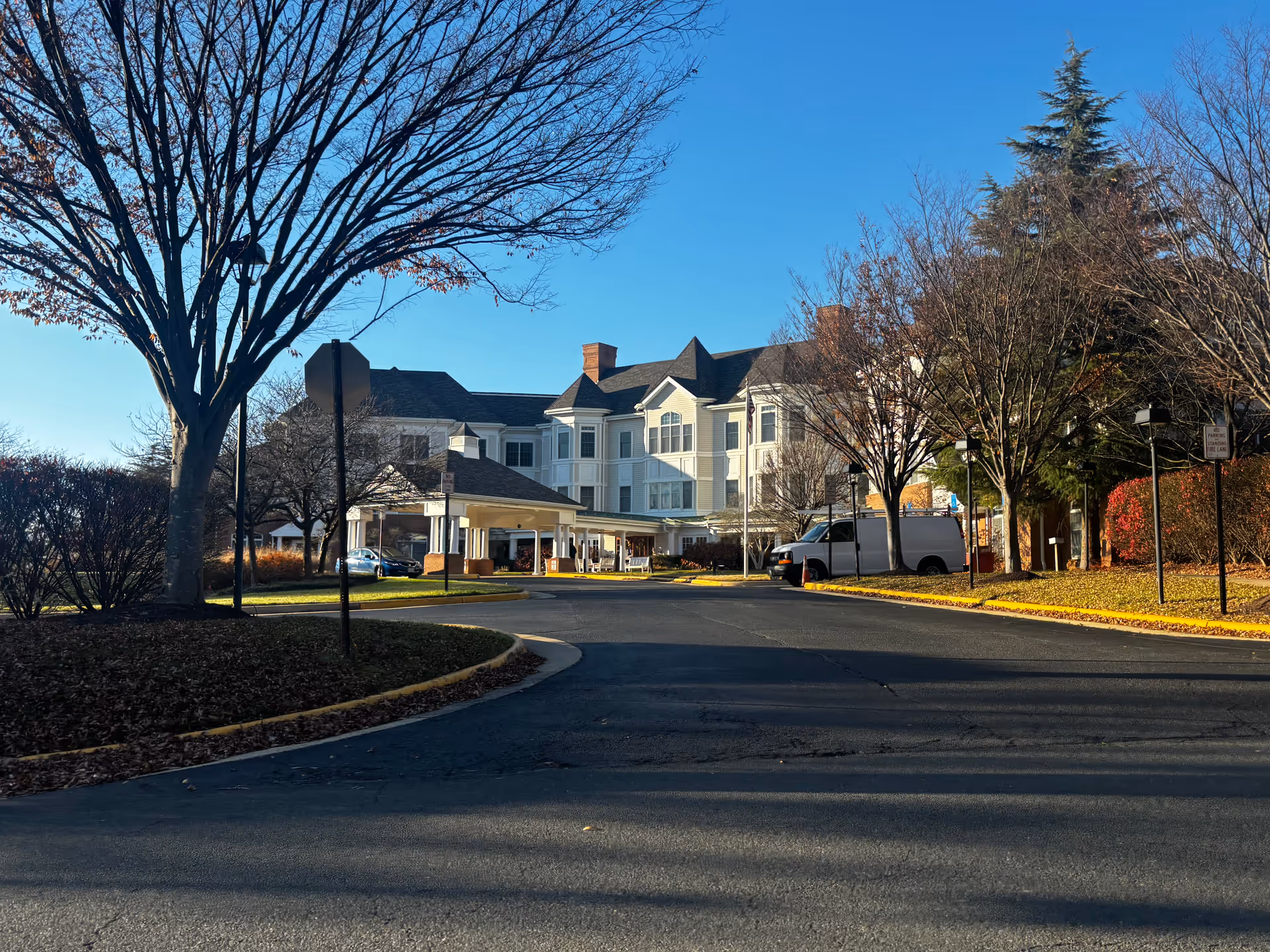 Exterior view of a large senior living facility building with white siding and multiple windows, surrounded by leafless trees and a clear blue sky. There is a driveway leading up to the entrance with a covered drop-off area, and a few vehicles parked nearby.