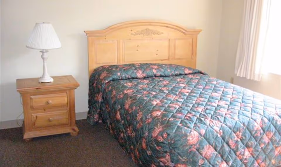 A simple bedroom with a wooden bed frame and headboard, a floral patterned quilt on the bed, a wooden nightstand with two drawers, and a white table lamp. There is a window with white curtains allowing natural light into the room.