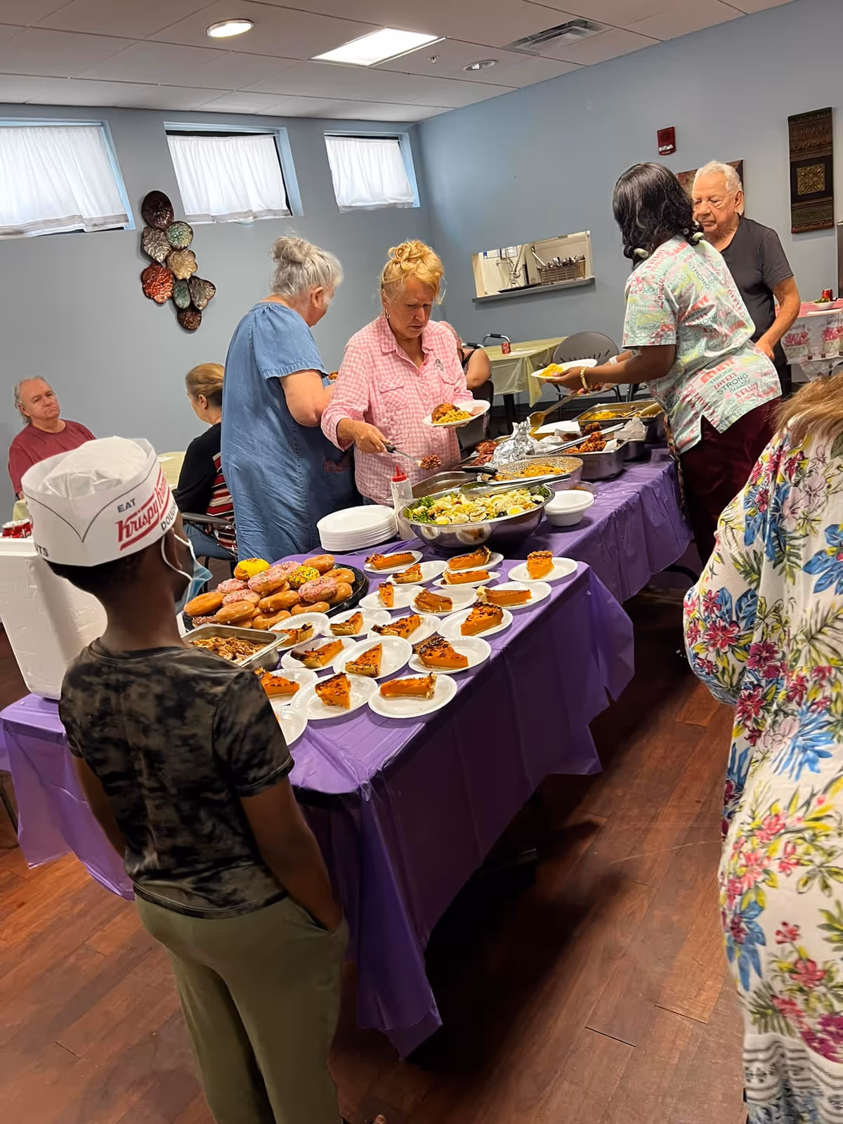 A group of people serving themselves food from a buffet table covered with a purple tablecloth in a room with light blue walls and three small windows. The buffet includes plates with slices of pie, a bowl of salad, and trays of hot food. A child wearing a Krispy Kreme paper hat stands near the table, while several elderly individuals are in line or seated in the background.