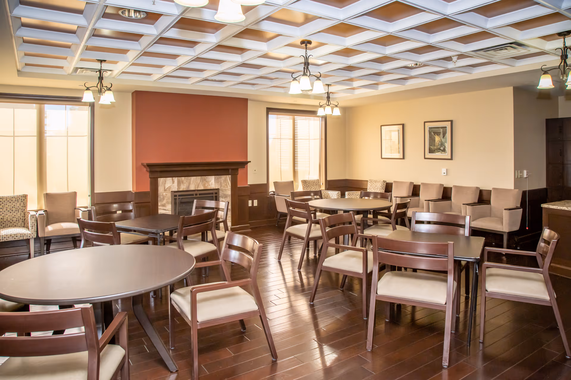 A well-lit room with multiple round and rectangular tables surrounded by wooden chairs with beige cushions. The room features a fireplace with a red accent wall behind it, large windows with blinds, framed artwork on the walls, and a coffered ceiling with hanging light fixtures. The floor is dark hardwood, and there are additional cushioned chairs along the walls.
