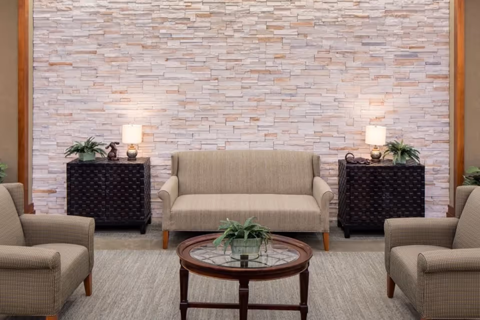 A cozy seating area with a beige loveseat centered against a textured stone wall, flanked by two dark woven side tables each holding a small lamp and a plant. Two matching beige armchairs face the loveseat with a round wooden coffee table with a glass top and a small plant in the center.